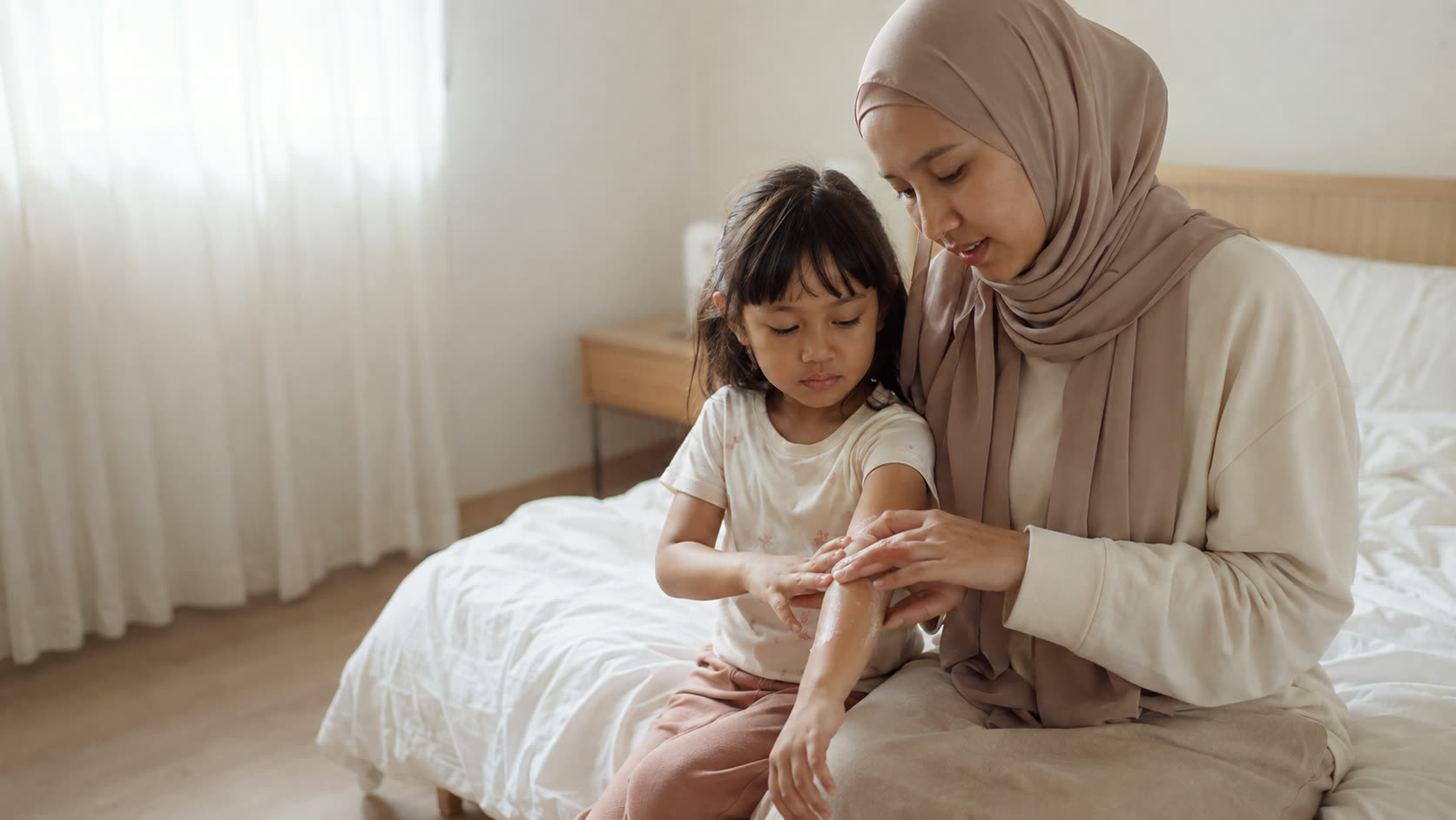 Mother applying moisturiser to her young daughter's arm in a sunlit Malaysian bedroom