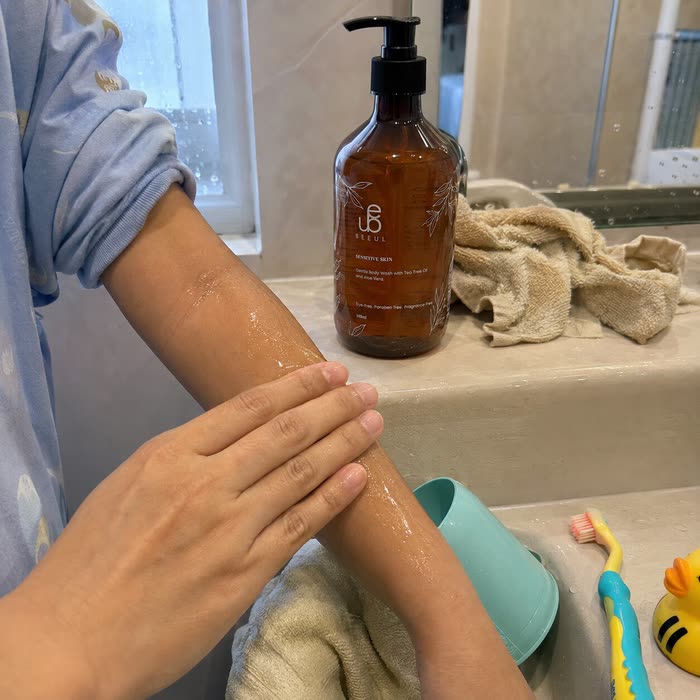 A child's forearm being washed at a bathroom basin, the BEEUL Gentle Body Wash bottle on the ledge behind