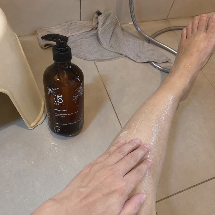 BEEUL Gentle Body Wash on a bathroom floor beside a stool, a hand smoothing water along a wet shin